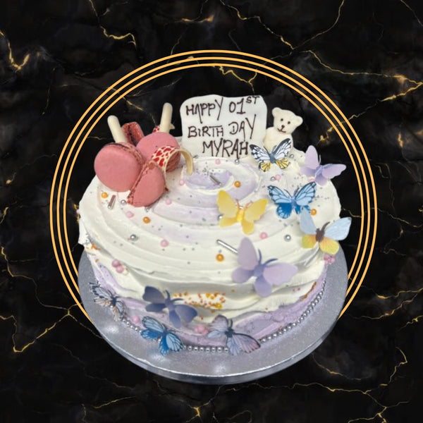 White and pastel birthday cake decorated with butterflies, pink macarons, and a teddy bear topper